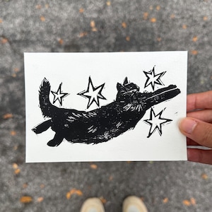May include: A black and white linocut print of a cat leaping through the air, surrounded by stars. The cat is silhouetted against a white background. The print is rectangular and held by a hand.