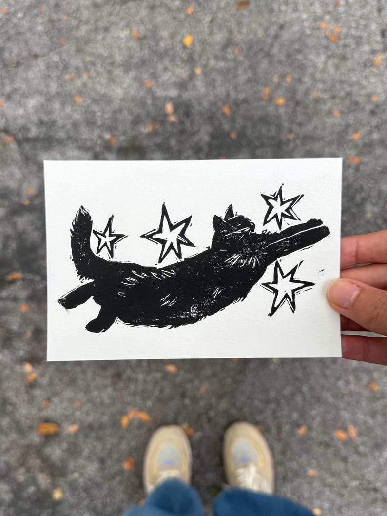 May include: A black and white linocut print of a cat leaping through the air, surrounded by stars. The cat is silhouetted against a white background. The print is rectangular and held by a hand.