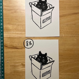 May include: Two rectangular white paper prints featuring a black ink linocut illustration of a cat sitting inside a cardboard box. The top print is labeled 82 and the bottom print is labeled 83. A green ruler is visible on the left side.