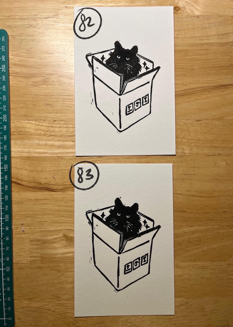 May include: Two rectangular white paper prints featuring a black ink linocut illustration of a cat sitting inside a cardboard box. The top print is labeled 82 and the bottom print is labeled 83. A green ruler is visible on the left side.