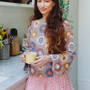 Könnte beinhalten: Eine Frau trägt einen rosa-braunen Häkelpullover mit einem Rock mit Blumenmuster und hält eine gelbe Tasse in ihren Händen. Sie steht in einer Küche mit einem Regal hinter ihr.