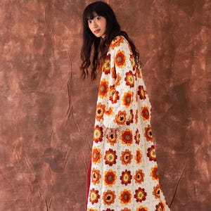 Puede incluir: Un cárdigan largo de crochet blanco con un patrón floral en tonos naranja, amarillo y rojo. El cárdigan se lleva sobre una falda roja y zuecos marrones.