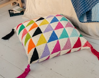 May include: A colorful crocheted floor pillow with a geometric pattern of triangles in various colors, including pink, blue, green, yellow, orange, and black. The pillow has two pink tassels.