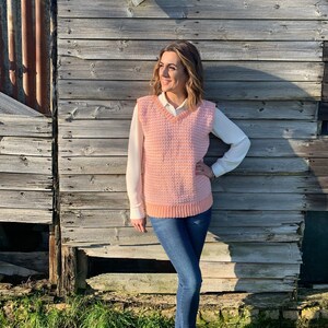 May include: A light pink knitted vest with a V-neck. The vest is worn over a white long-sleeved shirt and paired with blue jeans and brown ankle boots. The background is a weathered wooden wall.
