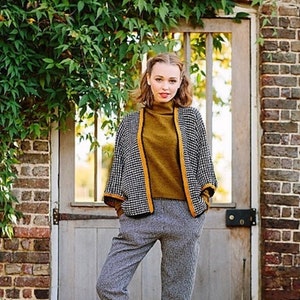 May include: A woman wearing a black and white checkered jacket with yellow trim, a brown turtleneck sweater, and gray checkered pants. She is standing in front of a wooden door with a brick wall behind her. There are fallen leaves on the ground.
