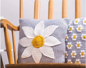 May include: Two crocheted throw pillows with a daisy design. One pillow features a large white daisy with a yellow center, while the other pillow has a pattern of smaller white daisies with yellow centers on a gray background.