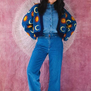Könnte beinhalten: Eine Frau trägt eine blaue Häkeljacke mit gelben Sonnen- und Mondmotiven, ein Jeanshemd und blaue Jeans. Die Jacke hat ein himmlisches Muster. Das Outfit ist lässig und stilvoll.