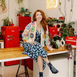 May include: A woman wearing a floral crochet cardigan and a blue floral dress sits on a wooden counter in a studio. The studio has red appliances and a neon sign that reads "Peanut Factory Studio".