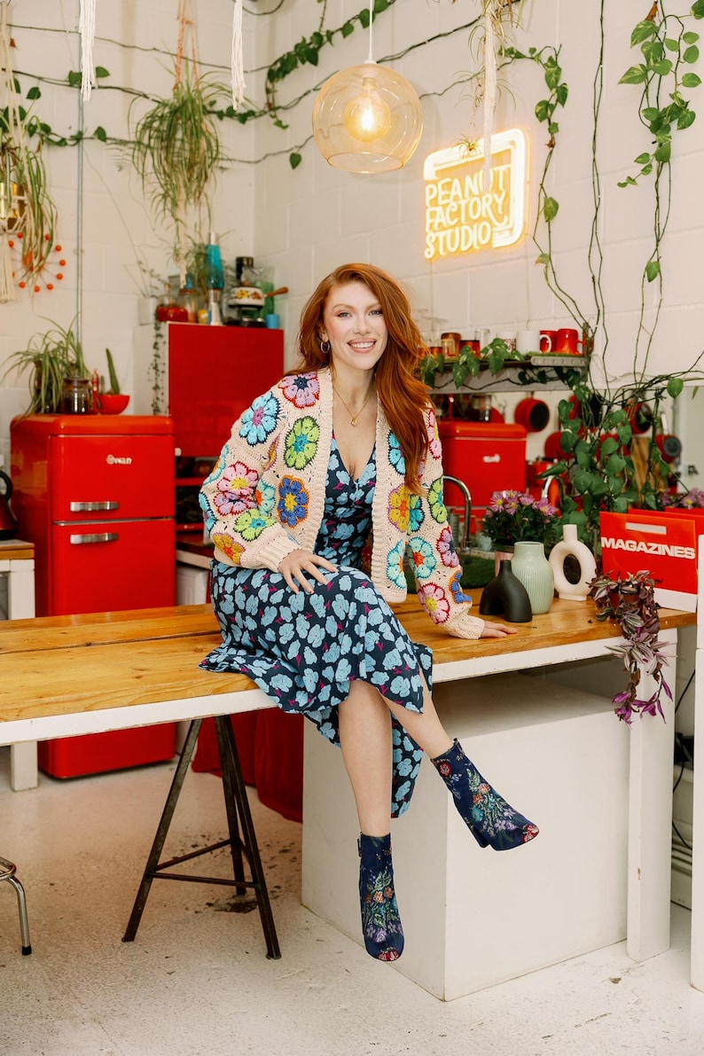 May include: A woman wearing a floral crochet cardigan and a blue floral dress sits on a wooden counter in a studio. The studio has red appliances and a neon sign that reads "Peanut Factory Studio".