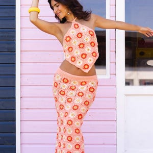 May include: A peach crochet halter top and skirt set with a floral pattern in white and red. The outfit is worn with platform sandals and a yellow bracelet. The background is a pink building. The outfit is a summer style.