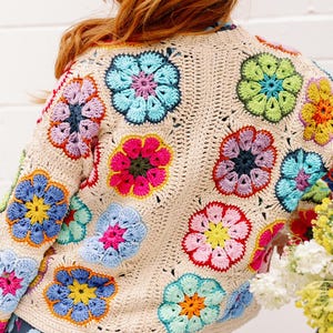 May include: A crocheted cardigan with a floral design in shades of pink, blue, green, and yellow. The cardigan is made with a beige yarn and has a loose fit.