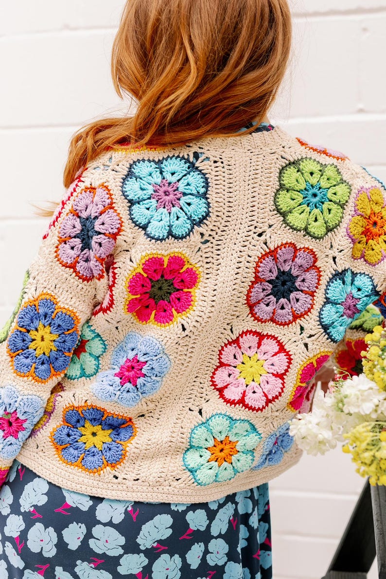 May include: A crocheted cardigan with a floral design in shades of pink, blue, green, and yellow. The cardigan is made with a beige yarn and has a loose fit.