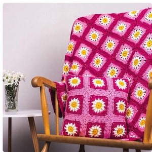 May include: A pink and white crocheted throw blanket with a daisy pattern is draped over a wooden rocking chair. A matching pink and white crocheted pillow with a daisy pattern is on the chair.