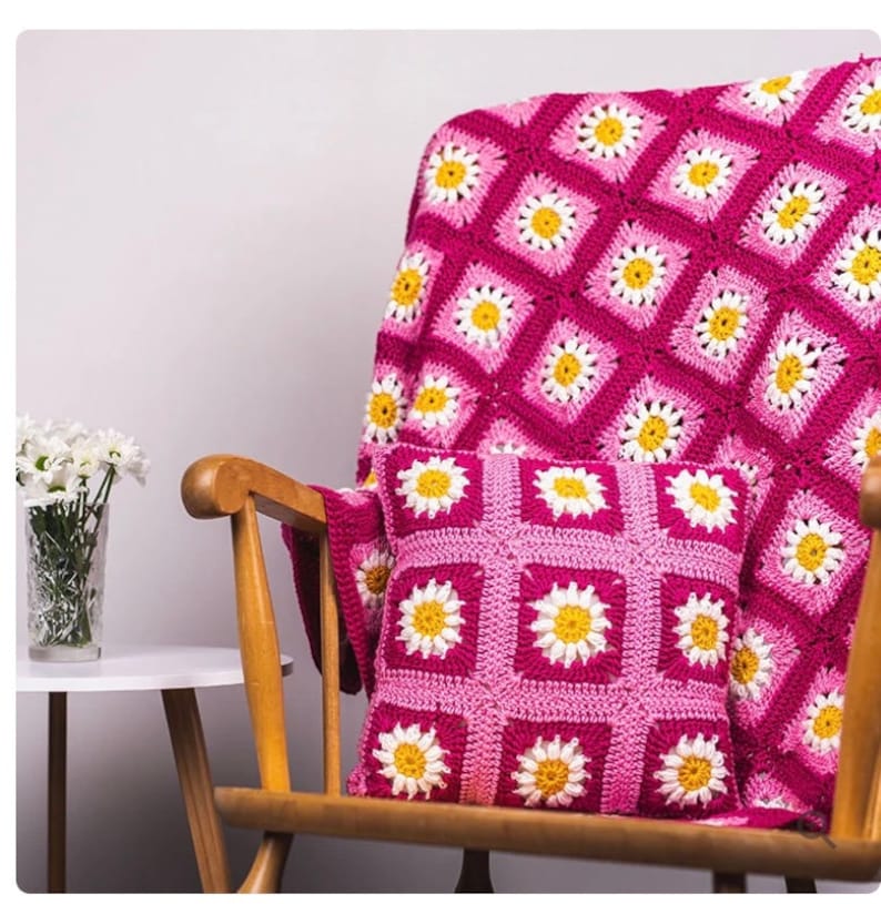 May include: A pink and white crocheted throw blanket with a daisy pattern is draped over a wooden rocking chair. A matching pink and white crocheted pillow with a daisy pattern is on the chair.