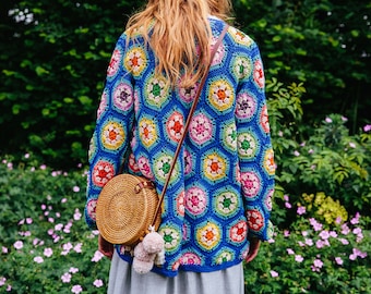 May include: A woman wearing a colorful crocheted cardigan with a round woven bag slung over her shoulder. The cardigan features a geometric pattern of hexagons in shades of blue, pink, yellow, green, and white.