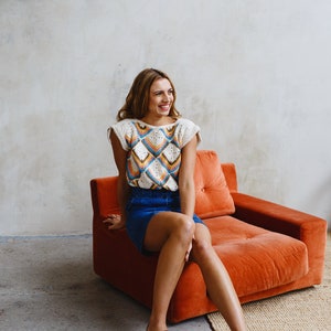 May include: A woman wearing a white crochet top with a colorful geometric pattern, a blue denim skirt, and brown sandals sits on an orange couch.
