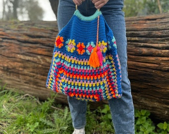 May include: A colorful crocheted tote bag with a blue base and a rainbow pattern. The bag has a gold metal handle and a bright orange tassel.