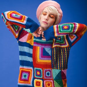 Op de afbeelding: Een kleurrijke patchwork gehaakte trui met een blauwe basis en levendige vierkanten van rood, oranje, geel, groen en paars. De trui heeft lange mouwen en een ronde hals. Een roze baret wordt op het hoofd gedragen.