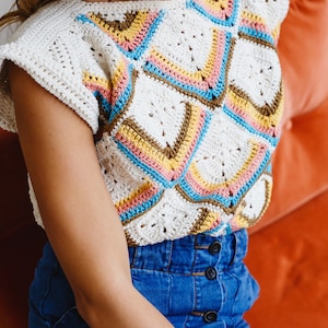 May include: A white crochet top with a colorful geometric pattern. The top has short sleeves and a round neckline. The model is wearing blue denim jeans.