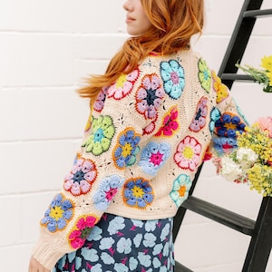 May include: A woman wearing a cream crochet cardigan with colorful flower motifs. The cardigan has a relaxed fit and long sleeves. The woman is wearing a blue floral skirt.