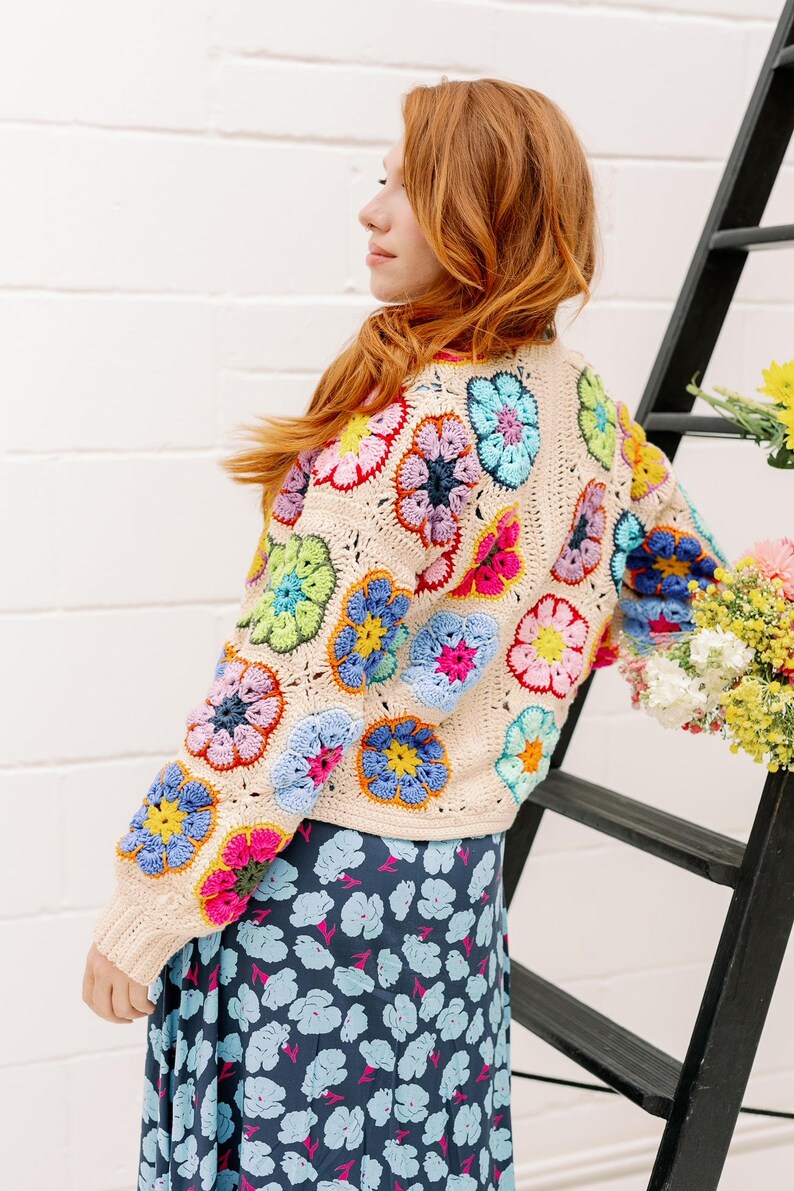 May include: A woman wearing a cream crochet cardigan with colorful flower motifs. The cardigan has a relaxed fit and long sleeves. The woman is wearing a blue floral skirt.
