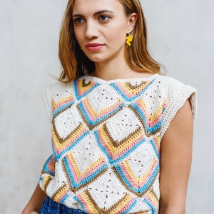 May include: A woman wearing a white crochet top with a colorful geometric pattern and blue denim jeans. She is wearing yellow earrings.