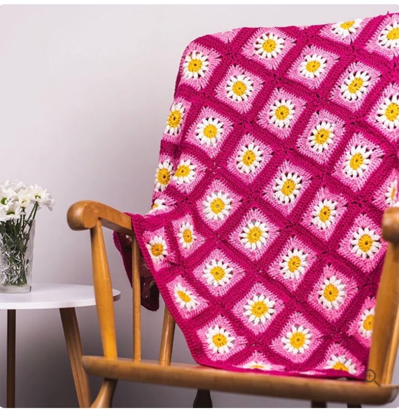May include: A pink and white crocheted throw blanket with a daisy pattern. The blanket is draped over a wooden rocking chair.