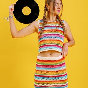 以下が含まれることがあります： カラフルなストライプのクロシェトップとそれに合わせたスカートを着た女性。彼女は左手に黒いビニールレコードを持っています。