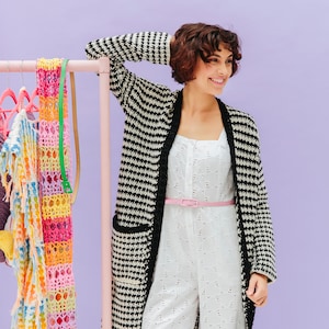 May include: A woman wearing a white jumpsuit with a black and white houndstooth cardigan. She is standing in front of a rack of colorful crocheted clothing.
