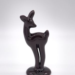 May include: A glossy, dark brown ceramic deer figurine. The deer is standing with its head turned, ears perked, and a slender body. The figurine is mounted on a circular base, showcasing a minimalist design.