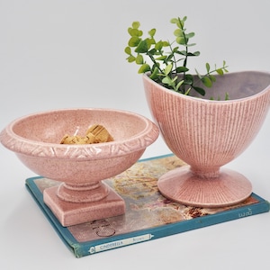 Könnte beinhalten: Zwei rosafarbene Keramik-Blumentöpfe mit gerippter Textur. Der größere Topf befindet sich links und hat einen breiten, runden Boden. Der kleinere Topf befindet sich rechts und hat einen hohen, schlanken Boden. Beide Töpfe stehen auf einem blauen Buch mit dem Wort "Cinderella" auf dem Einband.