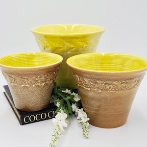 Puede incluir: Tres macetas de cerámica amarilla con un esmalte marrón y un diseño de hojas. Las macetas están dispuestas en forma de pirámide, con la maceta más grande en la parte superior y la más pequeña en la parte inferior. Las macetas están sobre un libro negro con el texto "Coco Chanel" visible.