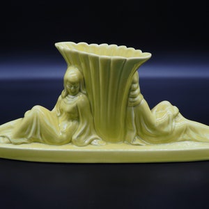 May include: A pale yellow ceramic vase with a central fluted design flanked by reclining female figures. The vase has a scalloped rim and sits on an elongated base. The figures are draped in flowing robes.