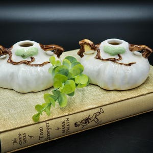 May include: Two white ceramic pumpkin-shaped candle holders with brown twig handles and green accents. The candle holders are sitting on a book titled "The Adventures of Tom Sawyer by Mark Twain".