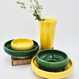 Puede incluir: Un juego de cuatro cuencos de cerámica amarilla y verde con un jarrón de cerámica amarillo con flores blancas. Los cuencos están apilados y tienen un interior verde. El jarrón es alto y delgado. Los cuencos están sobre un libro titulado "Las aventuras de Tom Sawyer por Mark Twain".