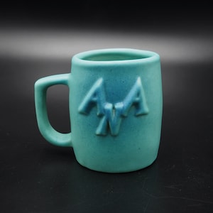 Puede incluir: Una taza de cerámica turquesa con un logotipo "ANA" en relieve. La taza tiene un cuerpo redondeado y un asa.