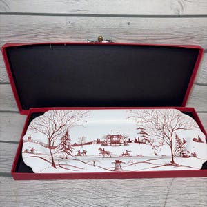 May include: A rectangular white ceramic serving platter featuring a detailed maroon winter landscape illustration with a large manor, bare trees, and figures on horseback. The platter is presented inside a red cardboard gift box with a black interior.