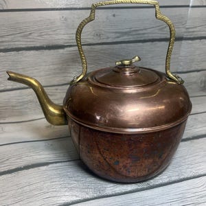 May include: A copper tea kettle featuring a polished brass spout and a textured brass handle. The kettle has a rounded body with a dark, weathered patina on the lower section and a smooth, shiny copper lid topped with a small brass bird finial.