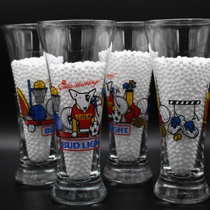 May include: Four clear glass Bud Light beer glasses with a white rim. Each glass features a different sports-themed design with a cartoon dog wearing a red, white, and blue jersey with the word "USA" on it. The text "Spuds MacKenzie" is printed on the glass.