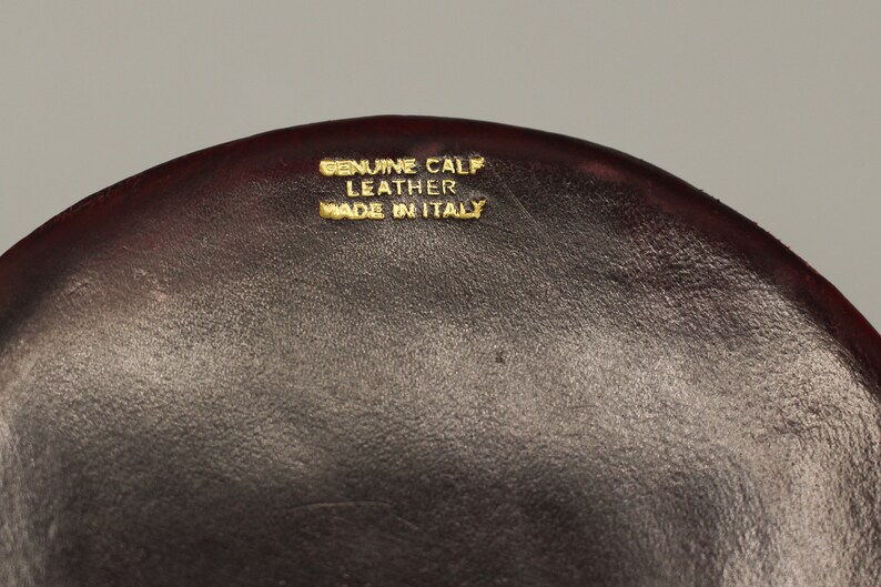 Może przedstawiać: Zbliżenie na ciemnobrązową sk&oacute;rzaną powierzchnię z złotym napisem "Genuine Calf Leather Made in Italy."