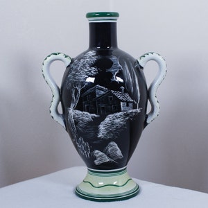 May include: A black ceramic vase with a landscape scene in white. The vase has two white handles with green accents, a green rim, and a green base. The landscape depicts a house, trees, and a path.