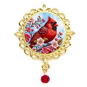 May include: A round gold-toned brooch featuring a vibrant red cardinal bird perched on a branch with pink blossoms against a light blue sky. The circular frame has an ornate filigree edge and a single dangling red faceted bead at the bottom.