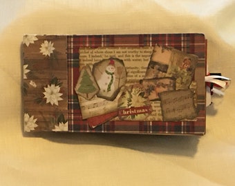 May include: A handcrafted Christmas journal with a wooden panel and poinsettia design. The cover has a plaid pattern and vintage-style images of a snowman, Christmas tree, and sheet music. Red and white ribbons are attached as a bookmark.