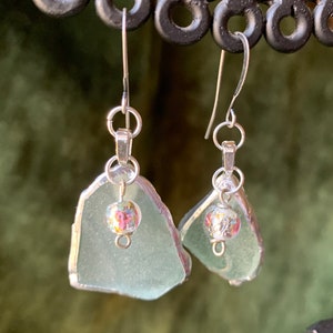 May include: A pair of handcrafted earrings featuring sea glass in a soft, pale green hue. Each earring has a silver-toned metal frame and a small, colorful bead with floral patterns. The earrings hang from silver-toned ear wires.