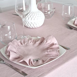 Op de afbeelding: Een roze linnen tafelkleed met een wit bord en een roze gekrulde servet. Er staan ​​verschillende heldere wijnglazen en een witte vaas met roze bloemen op de tafel.