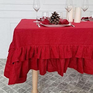 May include: Red linen tablecloth with a ruffled edge. The tablecloth is set on a table with a pine cone centerpiece and three wine glasses.