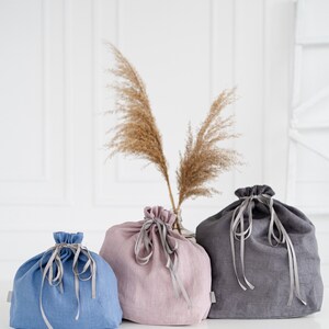 May include: Three linen drawstring bags in different colors: blue, pink, and gray. The bags are tied with gray ribbons and are sitting on a white surface.