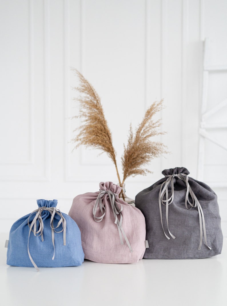 May include: Three linen drawstring bags in different colors: blue, pink, and gray. The bags are tied with gray ribbons and are sitting on a white surface.