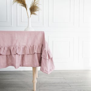 May include: A dusty rose linen tablecloth with a ruffled edge. The tablecloth is draped over a table with wooden legs. The table is set against a white wall with wainscoting.