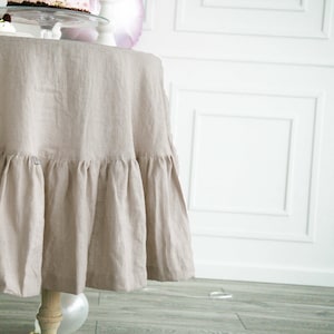 May include: A beige linen tablecloth with a ruffled edge. The tablecloth is draped over a round table and has a natural, rustic look.